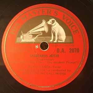 Pode incluir: Disco de vinil vermelho com um rótulo preto. O rótulo apresenta uma ilustração em preto e branco de um cão a ouvir um gramofone. O texto "His Master's Voice" está impresso a dourado sobre um fundo vermelho. O texto "D.A. 2070" está impresso a preto sobre um fundo branco. O texto "Gaudeamus Igitur (Traditional) Featured in the Film: "The Student Prince"" está impresso a preto sobre um fundo branco. O texto "Mario Lanza - Tenor Orchestra and Chorus conducted by Constantine Callinicos" está impresso a preto sobre um fundo branco.