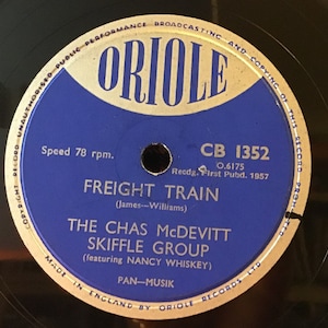 Può includere: Un disco in vinile blu con l'etichetta "ORIOLE" in lettere bianche. Il disco è intitolato "FREIGHT TRAIN" di "THE CHAS McDEVITT SKIFFLE GROUP" con la partecipazione di "NANCY WHISKEY". Il numero del disco è "CB 1352" e la velocità è di "78 giri al minuto".