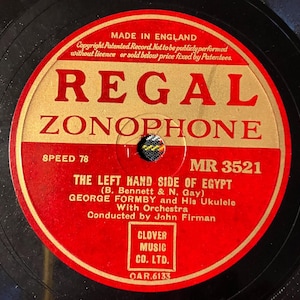 Puede incluir: Un disco Regal Zonophone vintage con una etiqueta roja y dorada. La etiqueta presenta el texto "THE LEFT HAND SIDE OF EGYPT" de George Formby and His Ukulele. El disco es de 78 RPM.