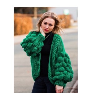 May include: A bright green, chunky knit cardigan with distinctive bubble sleeves. The open-front sweater is worn with a black top and dark trousers. The sleeves feature a textured, three-dimensional design.