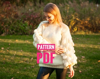 May include: Cream-colored, chunky knit sweater with textured bobble sleeves. The image displays the text "PATTERN PDF" in a pink box. The sweater is worn outdoors in a grassy area with a blurred background of trees.