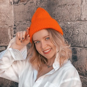 Peut inclure: Un bonnet en tricot orange vif est porté avec une chemise blanche boutonnée. Le bonnet est de couleur unie et contraste avec la chemise. La personne sourit, et l'arrière-plan est un mur de pierre texturé.