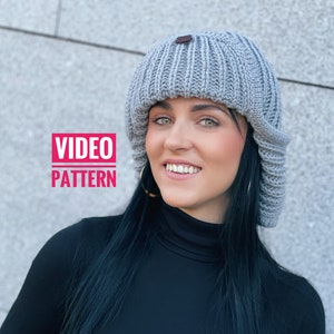 Peut inclure: Un bonnet en tricot côtelé gris clair avec une petite étiquette en cuir marron. Le bonnet est porté par une personne portant un col roulé noir. L'image comprend le texte "VIDEO PATTERN" dans un encadré rose.