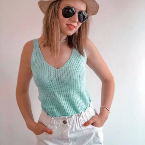 K&ouml;nnte beinhalten: Ein hellblaues Top mit V-Ausschnitt und offenem Gewebe wird mit einer wei&szlig;en Hose kombiniert. Das Outfit wird mit einem Strohhut mit schwarzem Band und einer schwarzen Sonnenbrille erg&auml;nzt. Die Person tr&auml;gt eine silberne Halskette und ein goldenes Armband.