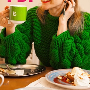 Könnte beinhalten: Eine Person trägt einen leuchtend grünen, grob gestrickten Pullover mit großen Ärmeln und hält eine grüne Tasse mit weißem Innenraum und dem Buchstaben "t". Ein Dessertteller und ein kleines Metalltablett stehen auf dem Tisch.