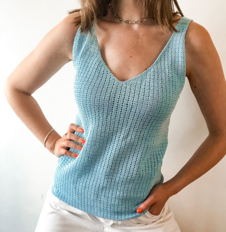 K&ouml;nnte beinhalten: Hellblaues &auml;rmelloses Tanktop mit V-Ausschnitt. Das Oberteil hat ein strukturiertes, offen gestricktes Design. Das Model tr&auml;gt eine silberne Halskette und ein silbernes Armband. Die Person tr&auml;gt wei&szlig;e Shorts. Der Hintergrund ist eine schlichte wei&szlig;e Wand.