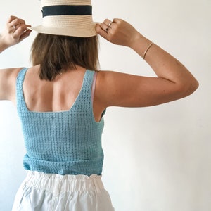 K&ouml;nnte beinhalten: Hellblaues H&auml;keltop mit quadratischem Ausschnitt und Strohhut mit schwarzem Band. Der Hut wird gehalten. Das Outfit wird durch wei&szlig;e Shorts erg&auml;nzt.