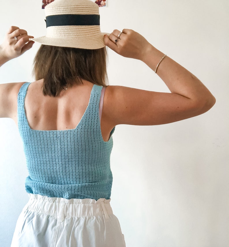 K&ouml;nnte beinhalten: Hellblaues H&auml;keltop mit quadratischem Ausschnitt und Strohhut mit schwarzem Band. Der Hut wird gehalten. Das Outfit wird durch wei&szlig;e Shorts erg&auml;nzt.