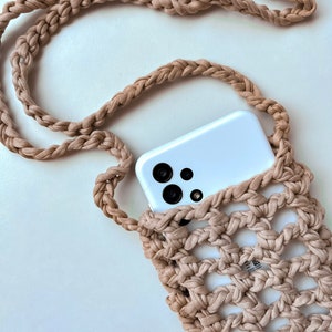 Op de afbeelding: Een beige macramé telefoon crossbody tas met een gevlochten riem. Het open geweven ontwerp bevat een witte smartphone. De tas is gemaakt van een zacht, geweven materiaal. De riem is lang genoeg om de tas crossbody te dragen.