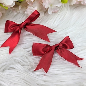 Könnte beinhalten: Zwei rote Satinbänder auf einer weißen, flauschigen Oberfläche. Die Schleifen sind mit einem Knoten in der Mitte gebunden und haben lange Bänder.