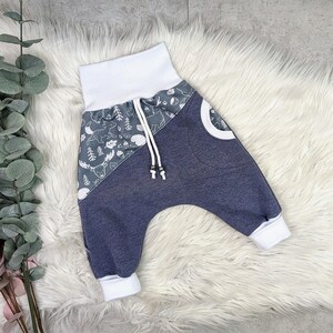 Könnte beinhalten: Blaue und graue Babyhose mit weißem Bund und weißem Saum an den Beinen. Die Hose hat auf der einen Seite einen Waldtierdruck und auf der anderen Seite eine einfarbige blaue Farbe.