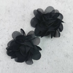 Könnte beinhalten: Zwei schwarze Stoffblumen-Verzierungen. Jede Blume besteht aus mehreren Schichten aus durchsichtigem schwarzem Stoff, was einen zarten und geschichteten Look erzeugt.