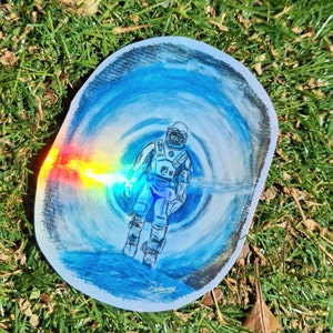 Puede incluir: Una pintura de acuarela de un astronauta con un traje espacial azul y blanco de pie en un vórtice azul y blanco giratorio. El astronauta sostiene un objeto brillante de colores arcoíris.