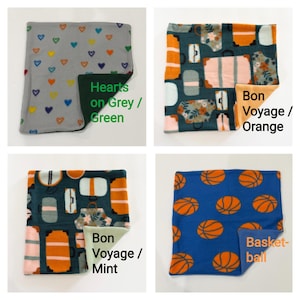 May include: Four square fleece fabric swatches. Top left: grey with multicolored hearts and dark green backing. Top right: travel luggage print with orange backing. Bottom left: travel luggage print with mint backing. Bottom right: blue with orange basketballs.