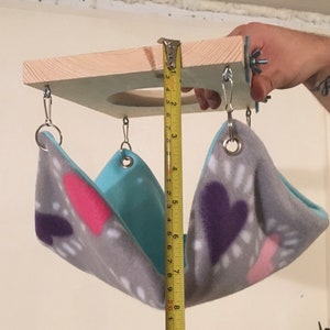 May include: A small animal hammock with a grey fleece fabric featuring pink and purple heart patterns and a solid light blue interior. The hammock hangs from a square wooden platform with metal hardware. A yellow measuring tape shows a depth of 8 inches.