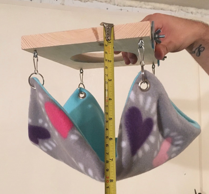 May include: A small animal hammock with a grey fleece fabric featuring pink and purple heart patterns and a solid light blue interior. The hammock hangs from a square wooden platform with metal hardware. A yellow measuring tape shows a depth of 8 inches.