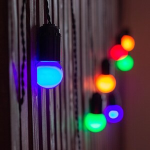 Pode incluir: Uma corda de lâmpadas LED coloridas penduradas em uma parede de madeira. As lâmpadas estão acesas em diferentes cores, incluindo azul, verde, vermelho e amarelo.