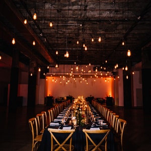 May include: A long, rectangular table with a black tablecloth is set for a formal dinner. The table is decorated with candles, greenery, and place settings. The table is surrounded by wooden chairs with cross-back designs. The room is dimly lit with string lights hanging from the ceiling.