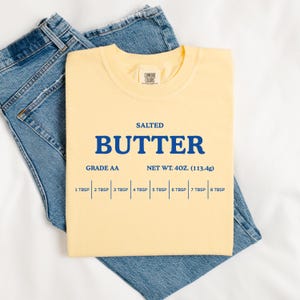 May include: A pale yellow crew neck t-shirt featuring blue text that reads SALTED BUTTER, GRADE AA, NET WT. 4OZ. (113.4g), and a measurement scale from 1 TBSP to 8 TBSP. The shirt is layered over a pair of blue denim jeans with visible stitching.