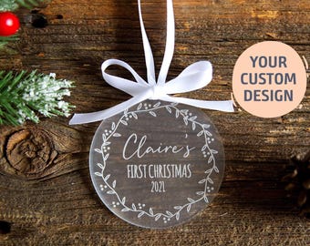 Puede incluir: Un adorno redondo de acrílico transparente con un diseño de corona impreso en blanco y el texto "Claire's FIRST CHRISTMAS 2021". El adorno está suspendido por una cinta de satén blanca atada en un lazo en la parte superior.
