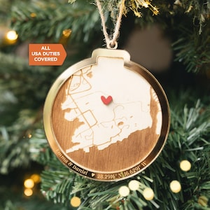 May include: A round, gold-rimmed Christmas ornament with a wooden map design. A small red heart marks a location on the map. The ornament has the text "Emma & Daniel" and coordinates. The text "ALL USA DUTIES COVERED" is also visible.