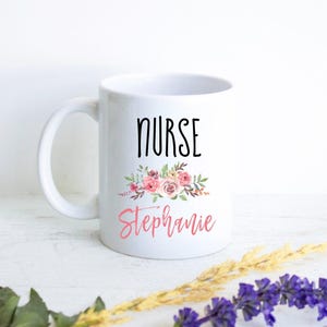Könnte beinhalten: Ein weißer Keramikbecher mit dem Wort NURSE in schwarzen Großbuchstaben über einer Blumenillustration aus rosa und violetten Rosen mit grünen Blättern, gefolgt von dem Namen Stephanie in einer rosa Schreibschrift.