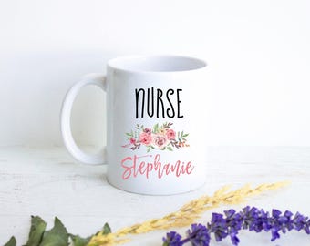 Könnte beinhalten: Ein weißer Keramikbecher mit dem Wort NURSE in schwarzen Großbuchstaben über einer Blumenillustration aus rosa und violetten Rosen mit grünen Blättern, gefolgt von dem Namen Stephanie in einer rosa Schreibschrift.