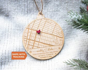 May include: A round wooden ornament featuring an engraved street map with a small red heart marker. The text reads: maddy & blake, First Christmas Our New Home, 40.7225 N, 73.9870 W, November 3, 2021. It hangs from a brown twine loop.