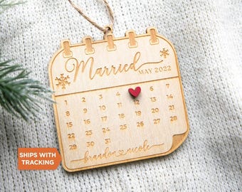 May include: A light-coloured wooden calendar ornament featuring the text Married MAY 2022, a small red heart marking the 12th, and the names Brandon and Nicole engraved at the bottom. The ornament includes snowflake icons and a brown twine hanging loop.