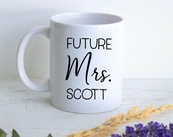 Könnte beinhalten: Ein weißer Keramikbecher mit dem schwarzen Aufdruck FUTURE Mrs. SCOTT. Der Becher hat eine standardmäßige zylindrische Form mit einem geschwungenen Henkel auf der linken Seite. Der Text ist in drei Zeilen angeordnet, wobei FUTURE in serifenloser Schrift.