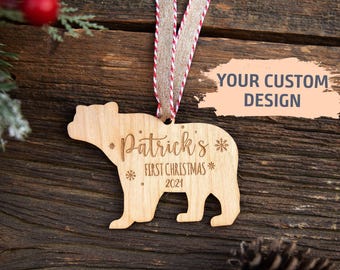 Op de afbeelding: Een lichtgekleurd houten ornament in de vorm van een beer met de gegraveerde tekst Patrick's First Christmas 2021 en sneeuwvlokken. Het ornament hangt aan een beige lint met dunne rode en witte strepen voor kerstdecoratie.