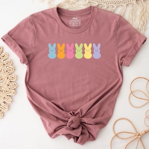 May include: A mauve short-sleeved crew neck t-shirt featuring a horizontal row of six stylised bunny silhouettes in pastel colours including blue, orange, pink, light green, yellow, and purple. The shirt has a standard fit with a knotted hem.
