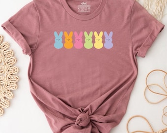 Könnte beinhalten: Ein malvenfarbenes Kurzarm-T-Shirt mit Rundhalsausschnitt, das eine horizontale Reihe von sechs stilisierten Hasensilhouetten in Pastellfarben zeigt, darunter Blau, Orange, Pink, Hellgrün, Gelb und Violett. Das Shirt hat eine klassische Passform mit einem geknoteten Saum.
