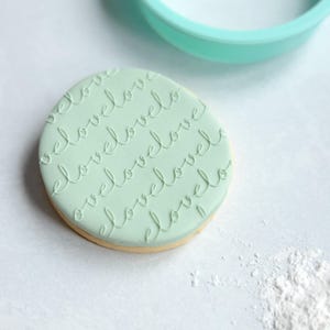 Può includere: Un biscotto rotondo ricoperto di pasta di zucchero liscia color verde menta pallido, caratterizzato da un motivo in rilievo ripetuto della parola love in corsivo. Un tagliabiscotti circolare color verde menta abbinato è posizionato parzialmente sopra il biscotto.