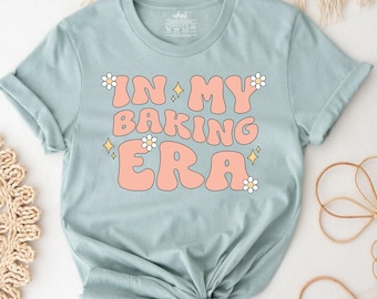 Peut inclure: Un t-shirt bleu clair à manches courtes avec le texte "IN MY BAKING ERA" écrit en grandes lettres arrondies de couleur pêche. Le motif est agrémenté de petites fleurs blanches de type marguerite et d'étoiles jaunes à quatre branches.