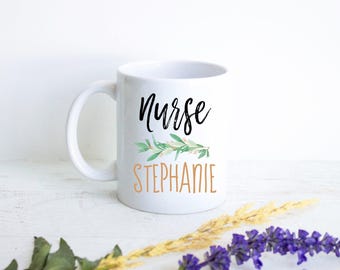May include: A white ceramic mug featuring the word Nurse in black cursive script above a green watercolor botanical sprig, with the name STEPHANIE printed in orange capital letters below the greenery on the front of the mug.