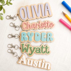 May include: Five wooden keychains with metal lobster clasps, each featuring a raised acrylic name in a different colour. The names displayed are OLIVIA in lavender, Charlotte in pink, RYDER in teal, Wyatt in sage green, and Austin in white.