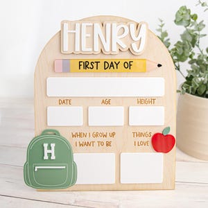 以下が含まれることがあります： 白い文字でHENRYと書かれたアーチ型の木製看板。FIRST DAY OFと記された黄色い鉛筆のグラフィックがあり、日付、年齢、身長、将来の夢、好きなものを記入するための白い長方形の空白スペースがある。緑色のリュックサックと赤いリンゴの装飾付き。
