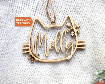May include: A light-colored wooden cat-shaped ornament featuring the name Molly in cursive script, accented with two snowflake designs on the sides and a brown string loop at the top for hanging, set against a textured white fabric background.