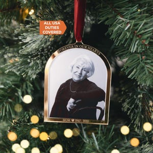 May include: A gold-toned metal arch-shaped memorial ornament featuring a black and white portrait of a person. The top is engraved with "FOREVER IN OUR HEARTS" and the bottom with "MOM 1945 - 2025". It hangs from a dark red ribbon.