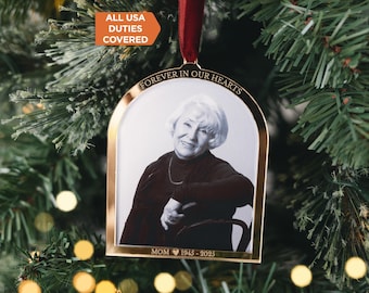 May include: A gold-toned metal arch-shaped ornament featuring a black and white portrait of a person. The top is engraved with the text FOREVER IN OUR HEARTS and the bottom with MOM 1945 - 2025. A dark red ribbon is attached to the top for hanging.