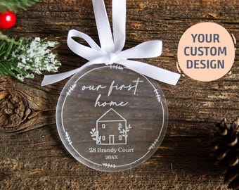 Pode incluir: Um ornamento circular de acrílico transparente com um laço de fita branca no topo. O ornamento está gravado com o texto "our first home", um desenho de linha de uma casa, "28 Brandy Court" e "20XX", rodeado por uma borda de folhas.