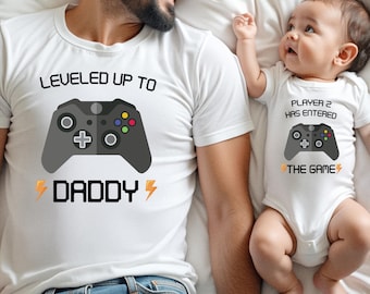 May include: A white t-shirt with a black video game controller graphic and the text LEVELED UP TO DADDY with orange lightning bolts, and a white baby bodysuit with a matching controller graphic and the text PLAYER 2 HAS ENTERED THE GAME.