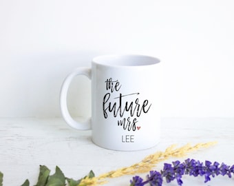 Könnte beinhalten: Ein weißer Keramikbecher mit Henkel, der den schwarzen Aufdruck "the future mrs." zusammen mit einem kleinen roten Herz und dem Namen "LEE" in schwarzen Großbuchstaben zeigt. Der Becher hat eine glatte Oberfläche und eine zylindrische Form.
