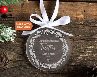 May include: A circular clear acrylic Christmas ornament featuring a white printed wreath design. The text reads Our First Christmas Together, EMILY & ALEX, and 2022, with a small heart icon. A white satin ribbon is tied in a bow at the top.