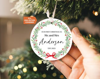 Puede incluir: Un adorno navideño redondo de cerámica blanca con una corona de acuarela verde, bayas rojas y un lazo rojo en la parte inferior. El texto dice: Our First Christmas as Mr. and Mrs. Anderson Est. 2023. Cuelga de una cinta blanca.