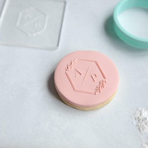 May include: A round biscuit topped with smooth pink fondant, embossed with a hexagonal frame, leaf accents, and the letters A and P separated by a diagonal line. A clear acrylic stamp and a teal circular biscuit cutter are positioned nearby.