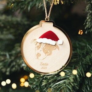 Peut inclure: Une décoration de Noël ronde en bois avec un bord doré, suspendue à une ficelle en corde naturelle. L'ornement présente un portrait de chien gravé au laser portant un bonnet de Père Noël. Le nom du chien, "Sadie", et l'année "2023" sont également gravés.