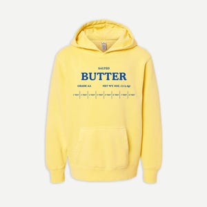 May include: A bright yellow hooded sweatshirt featuring a blue graphic print on the chest. The text reads SALTED BUTTER, GRADE AA, NET WT. 4OZ. (113.4g), and includes a measurement scale from 1 TBSP to 8 TBSP. The hoodie has a front kangaroo pocket.