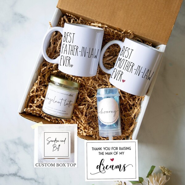 May include: A gift box containing two white ceramic mugs with black text reading Best Father-in-Law Ever and Best Mother-in-Law Ever, a jar candle labeled Hazelnut Latte, a confetti popper labeled Hooray, and a card reading Thank You For Raising The Man Of My Dreams.